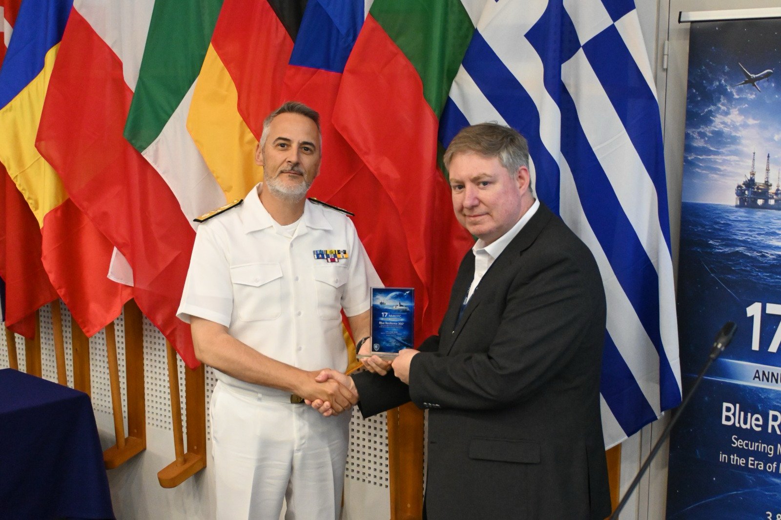 Adrian Neal receiving recognition at NMIOTC's annual NATO conference in Greece beside a naval officer and international flags